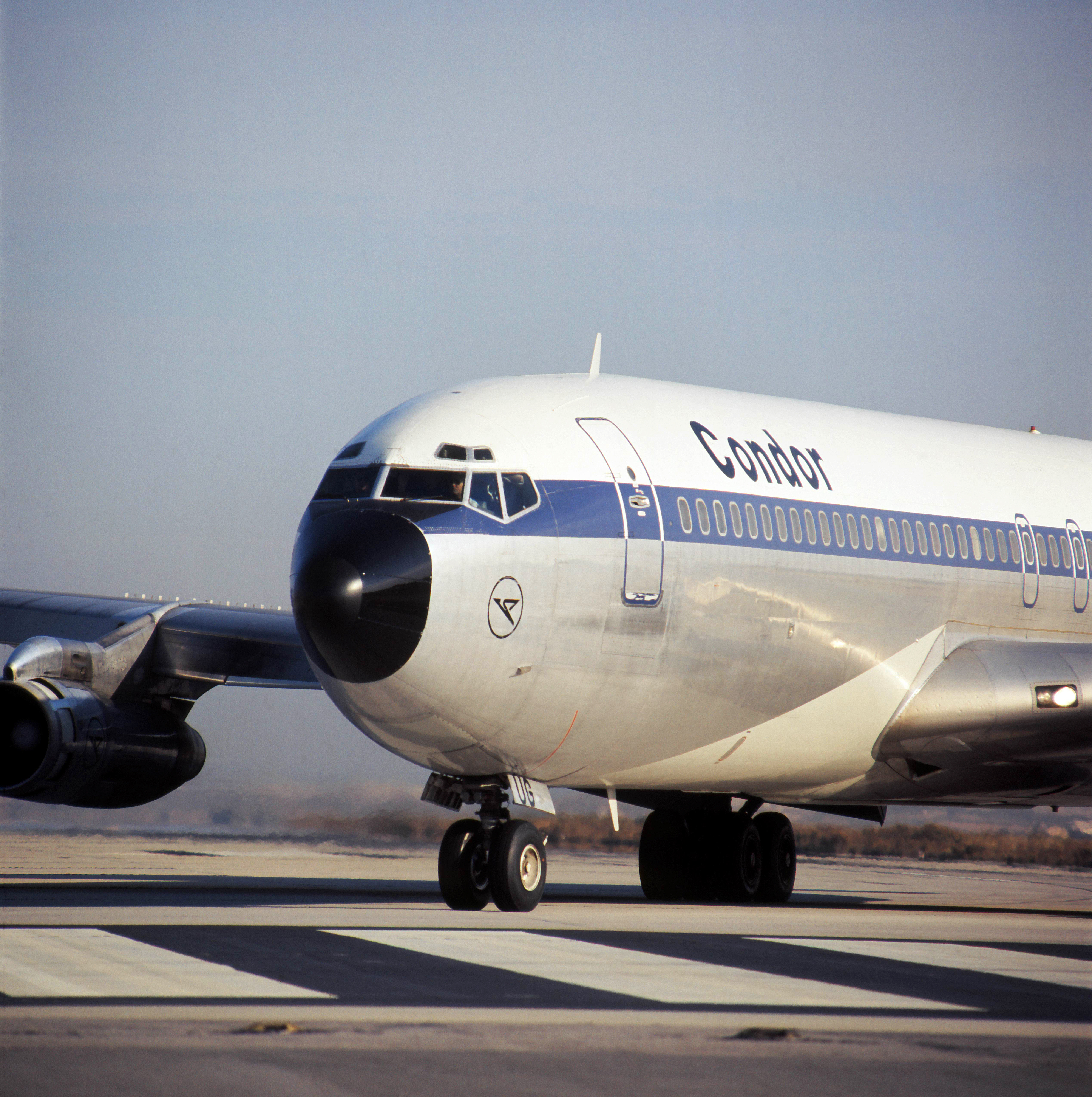 The image shows the nose of a vintage passenger airplane with blue-and-white livery on an airport apron. The cockpit windows, part of the aircraft fuselage and the engines are visible. The airplane is shown from the front and at an angle.