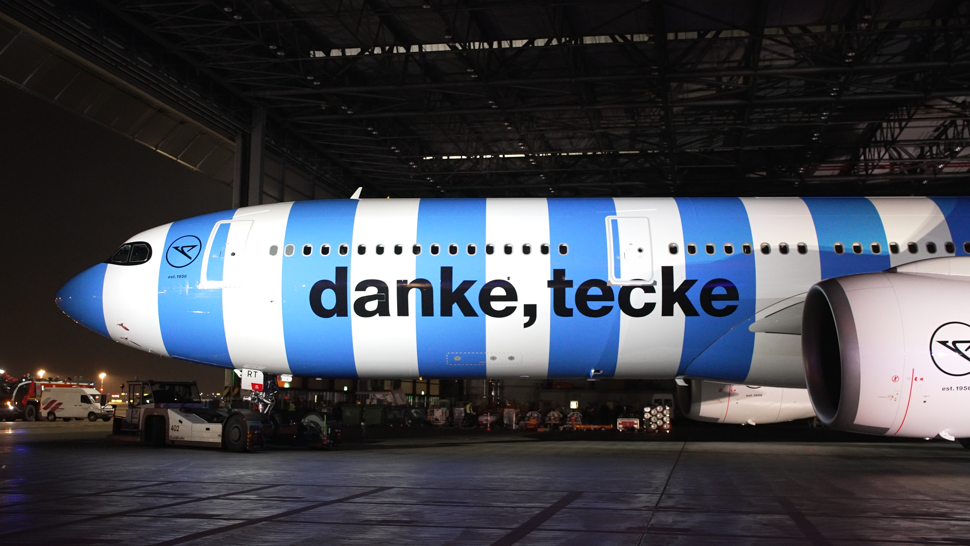 The image shows the fuselage of a passenger aircraft in blue-and-white-striped special livery in an aircraft hangar. The aircraft fuselage features the words “thank you, tecke.” The aircraft is shown in profile.